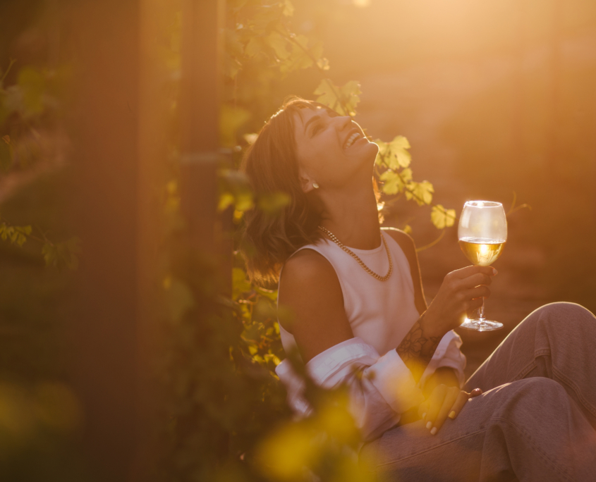 Young beautiful woman drinking wine in vineyard at sunset on summer sunny day.